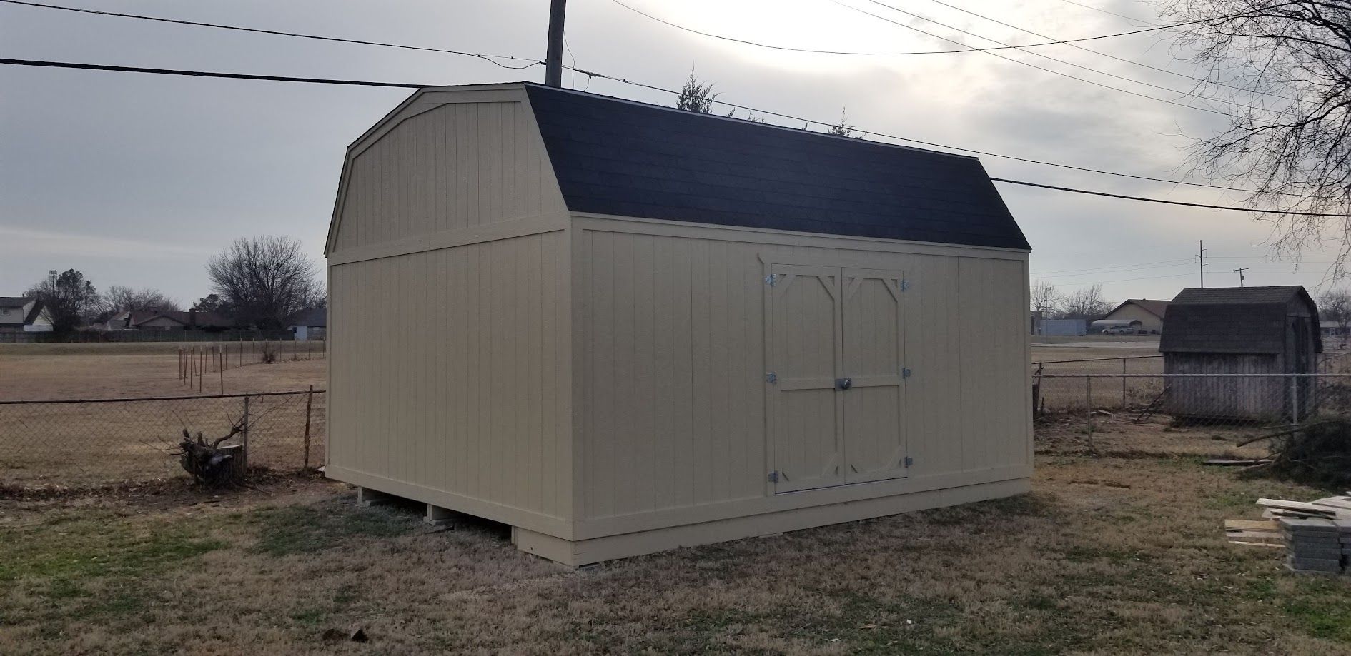 Beige shed with dark roof in a grassy rural yard under cloudy sky
