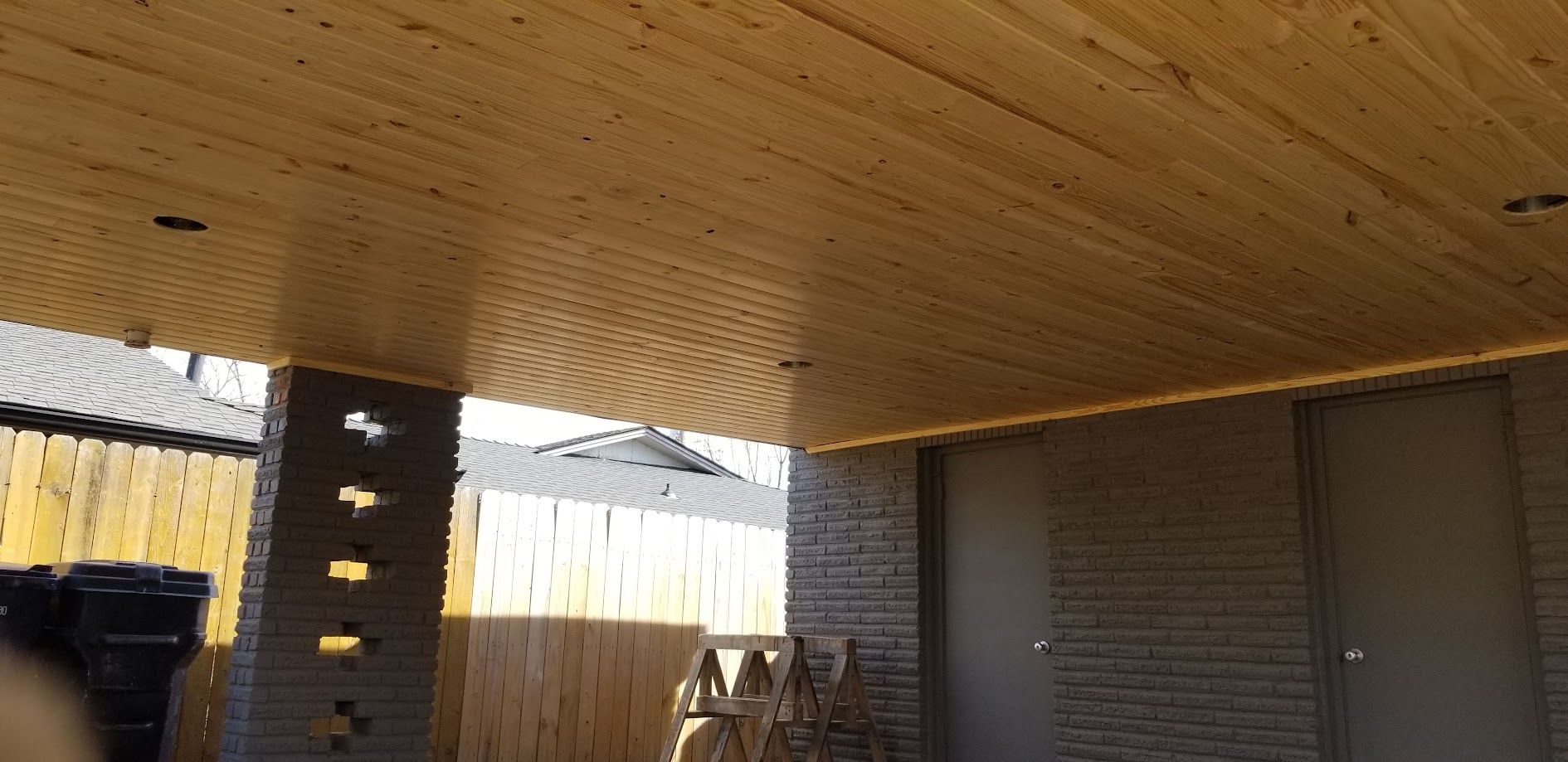 Wooden patio roof over brick and wood walls, with a fence and trees in the background.