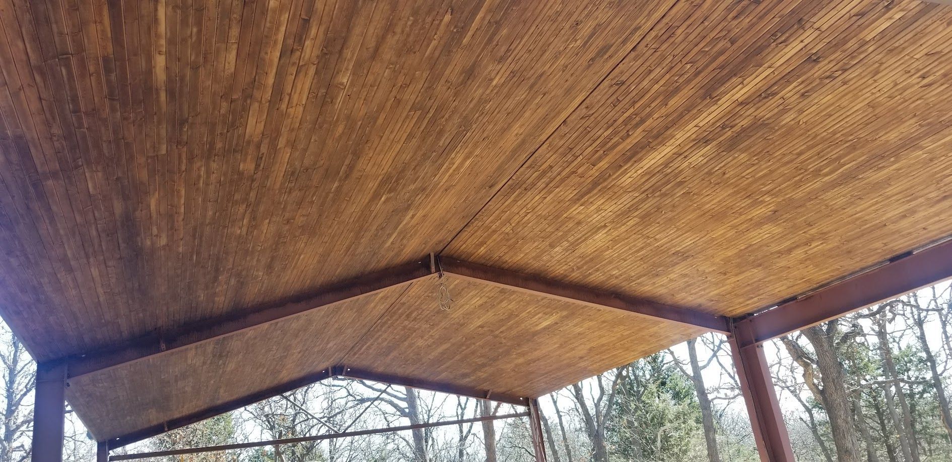 Wooden pavilion roof with red support posts, viewed from below against trees