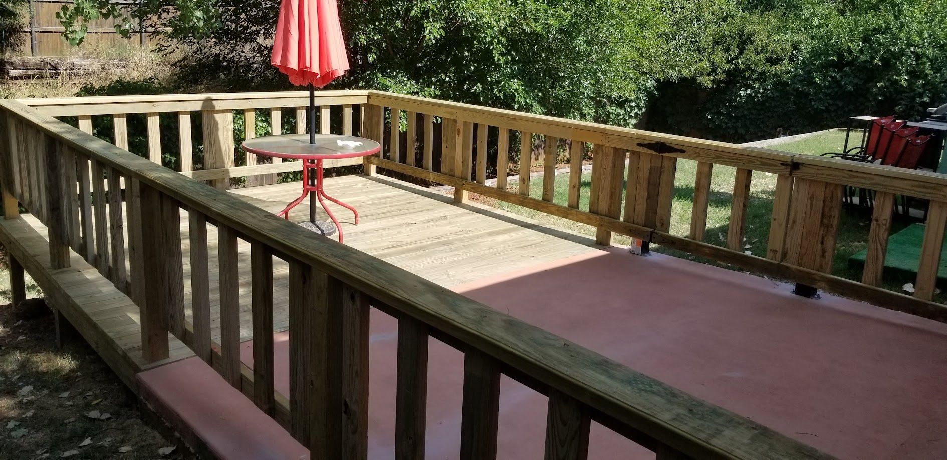 Wooden deck with railings and a red patio umbrella in a sunny wooded backyard