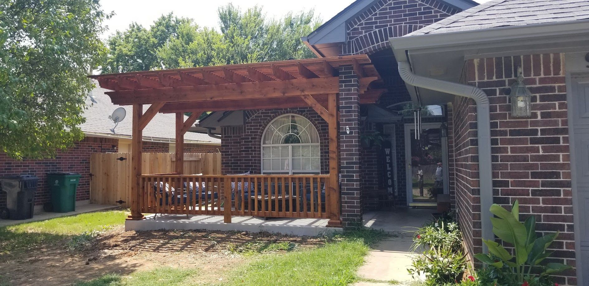 Brick house patio with a wooden pergola, railings, and a backyard fence in sunlight