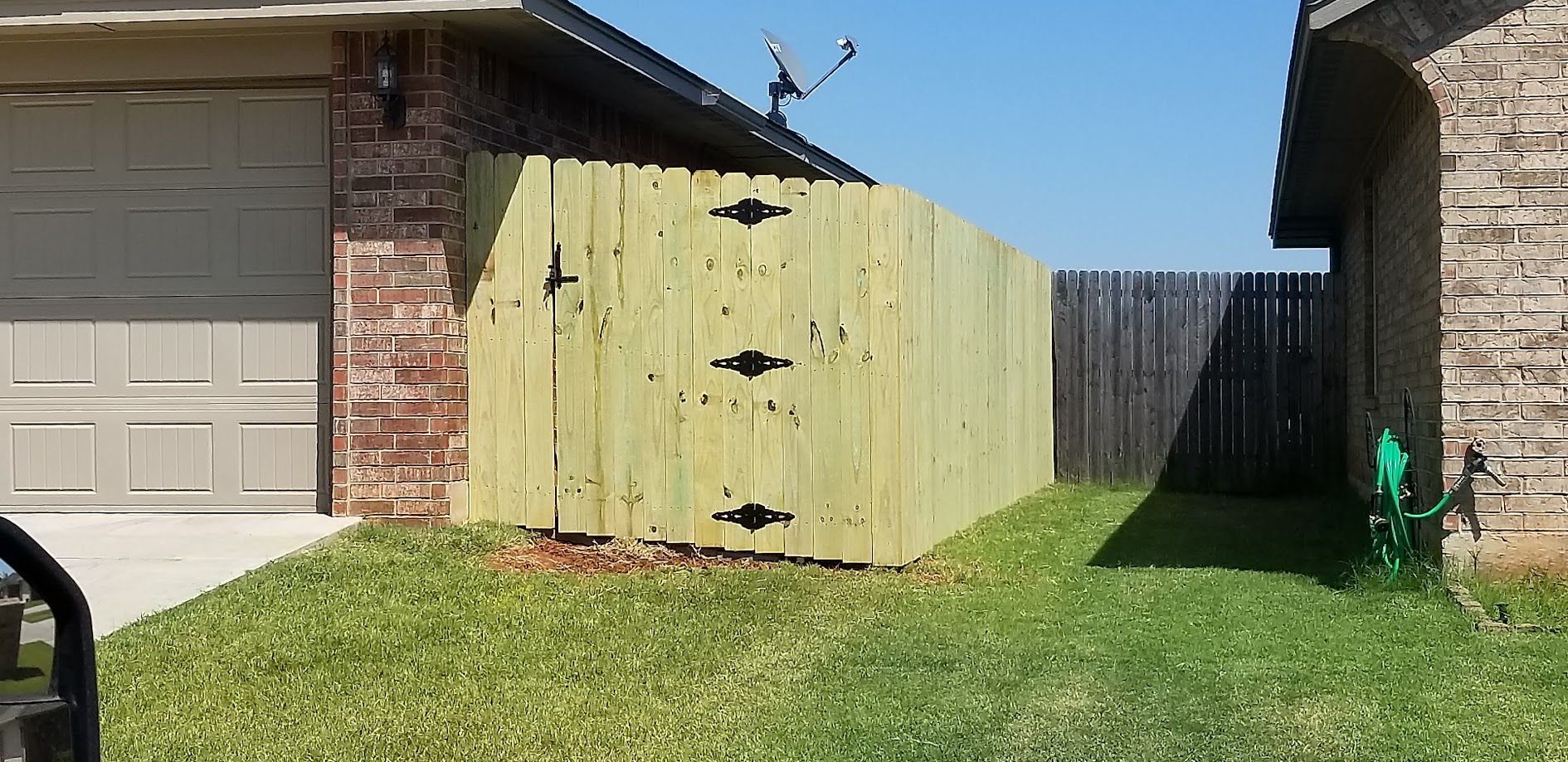 Green wooden fence beside a brick garage and grassy backyard under a clear blue sky