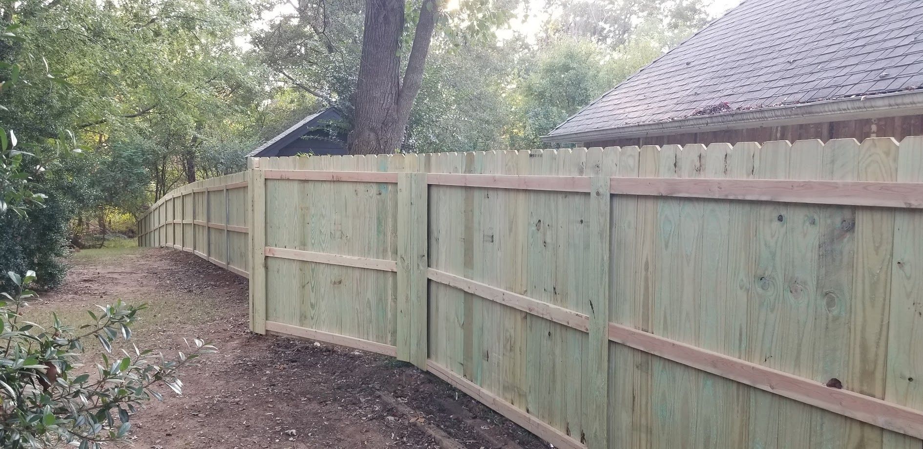 Wooden privacy fence along a dirt path beside trees and a house roofline