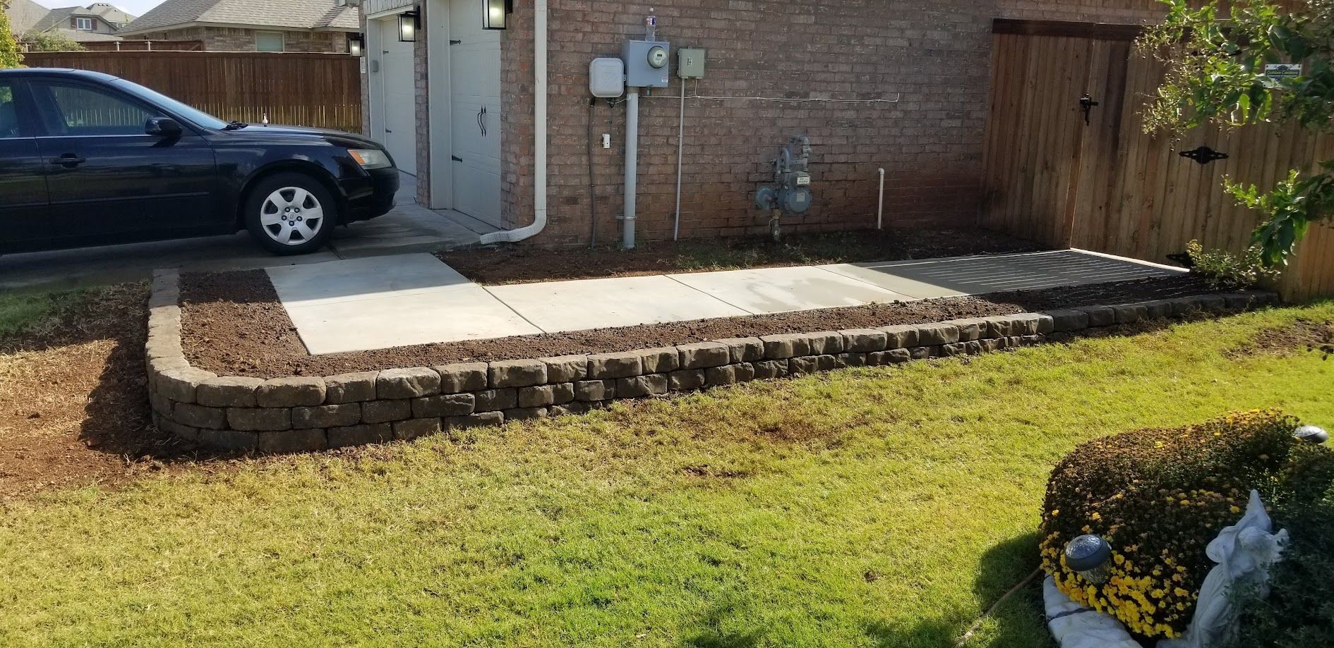 Raised concrete patio beside a brick house, with a black car parked nearby and a grassy yard in front.