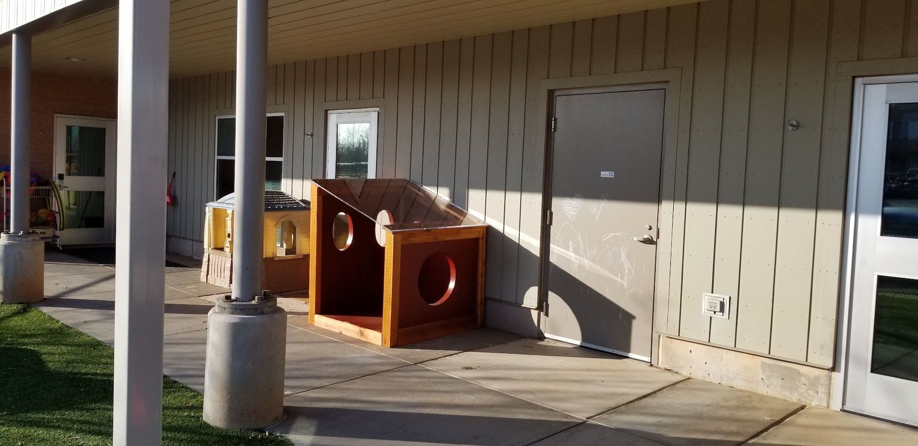 Red playground tunnel play structure beside a beige building under a covered walkway.