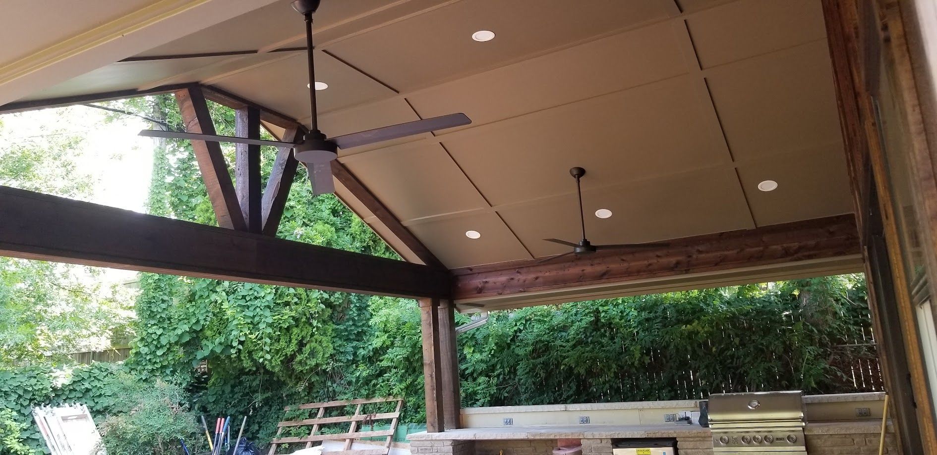 Covered outdoor patio with wooden beams, ceiling fans, and lush green trees beyond the railing