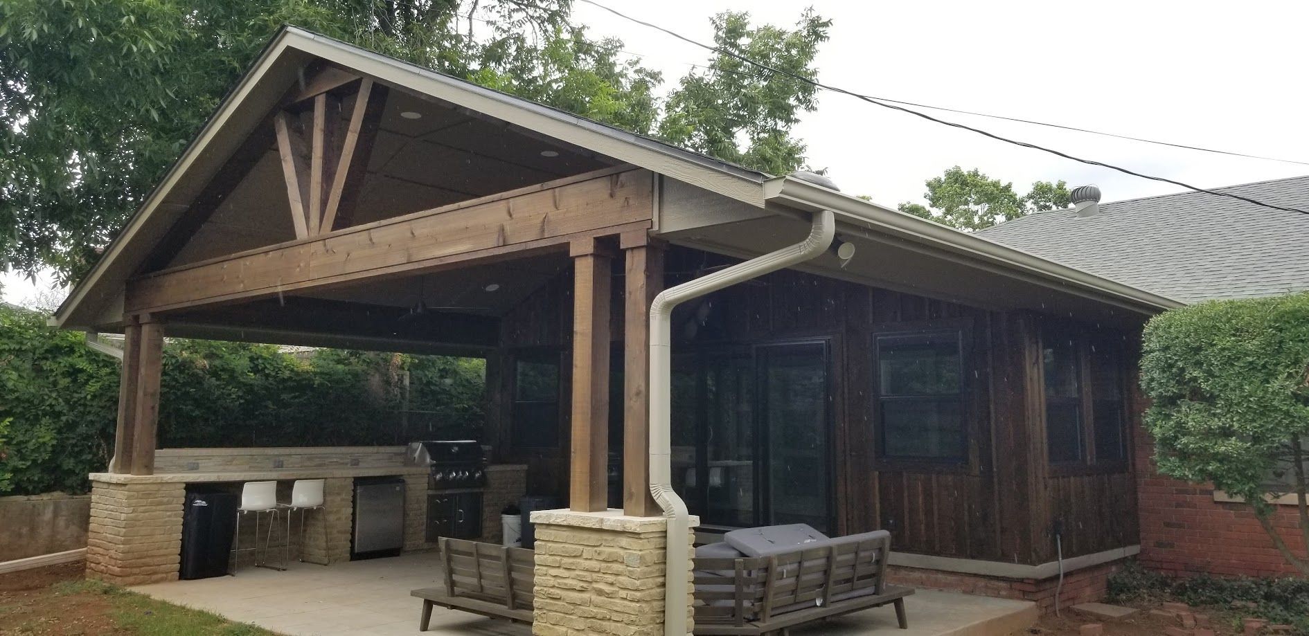Covered outdoor patio with wooden beams, stone pillars, and picnic tables beside a brick building