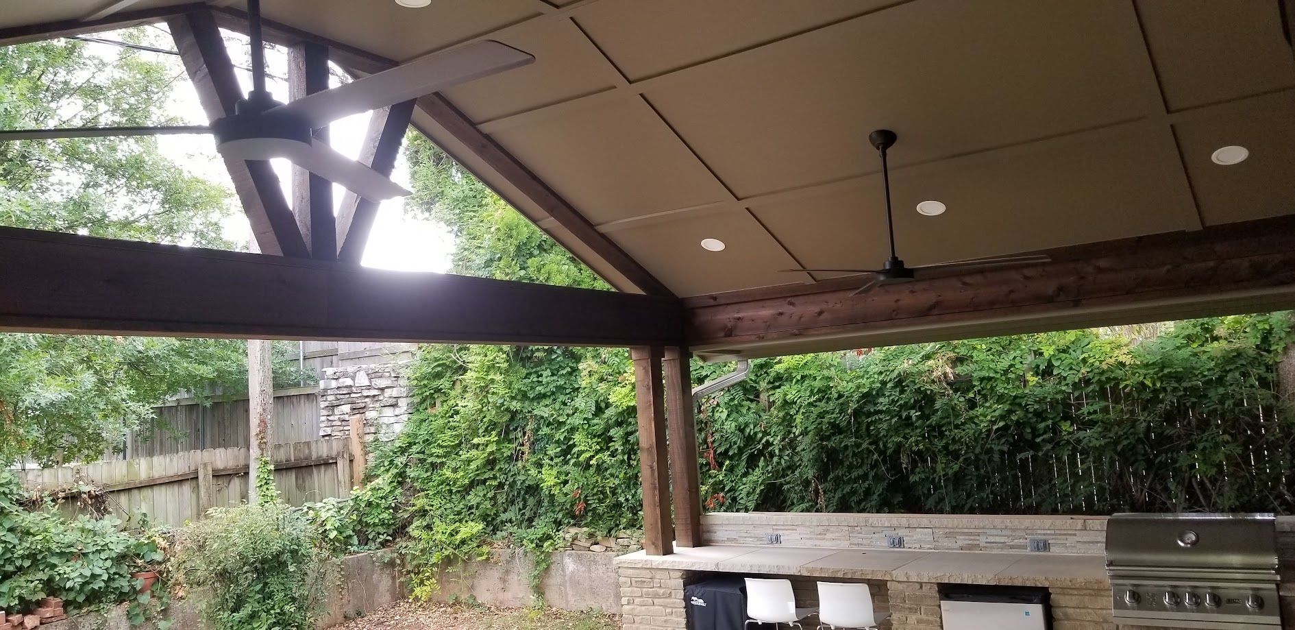 Covered outdoor patio with wooden benches, stone table, and greenery beyond a beam-framed roof.