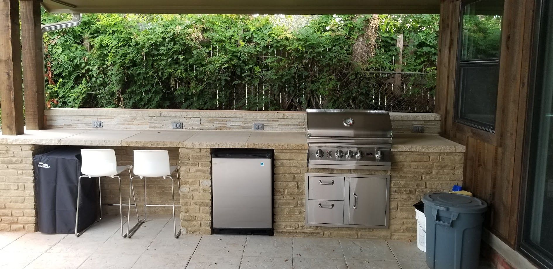 Outdoor patio kitchen with grill, mini fridge, sink, stools, and trash bins under a wooden roof