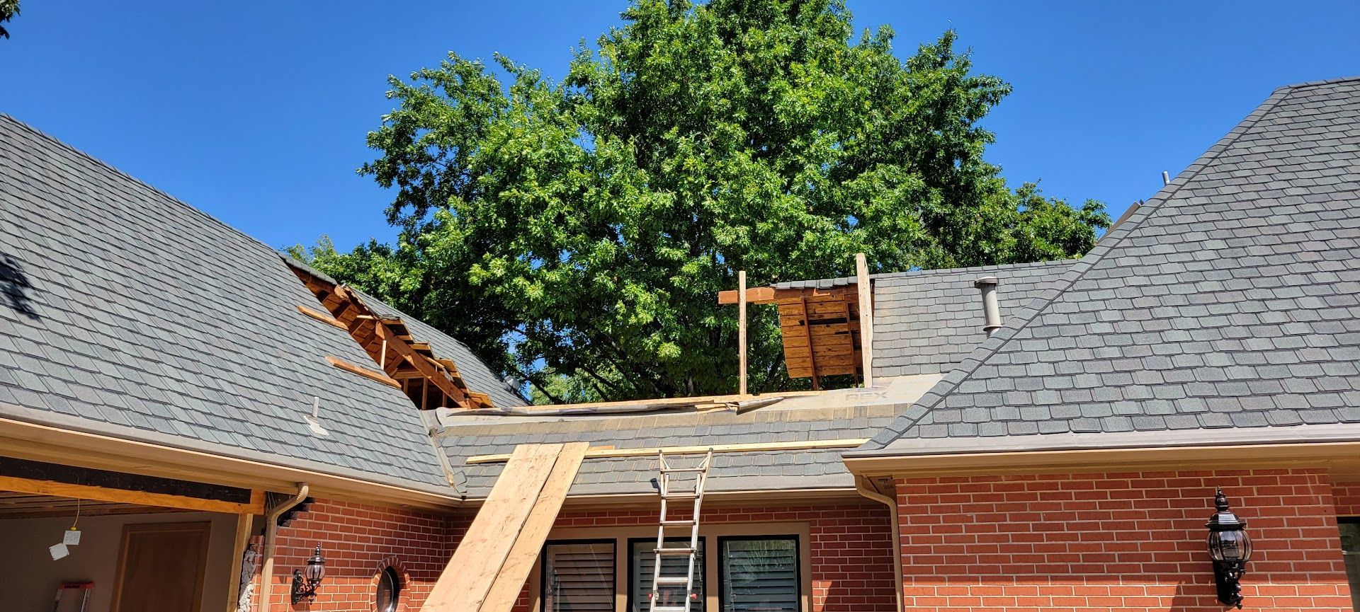 Roof repair on a brick house with a ladder and exposed decking between two gray shingled roof sections