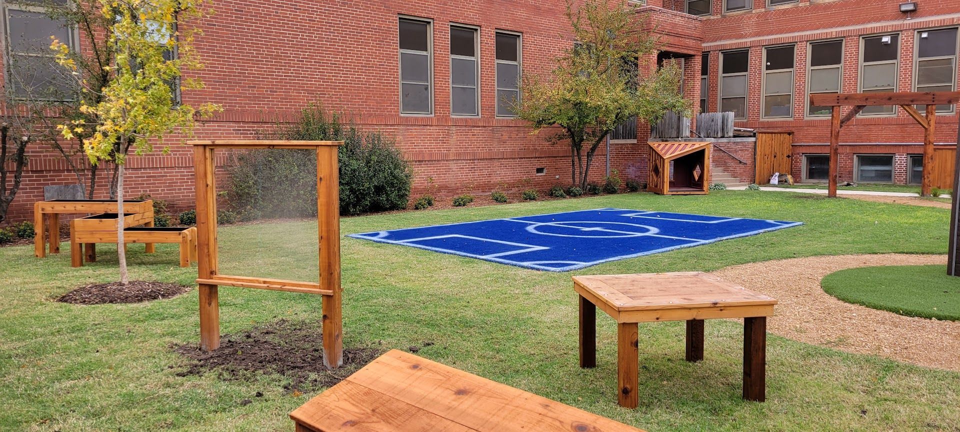 Outdoor courtyard with wooden structures, a blue mat, benches, grass, and a brick building backdrop