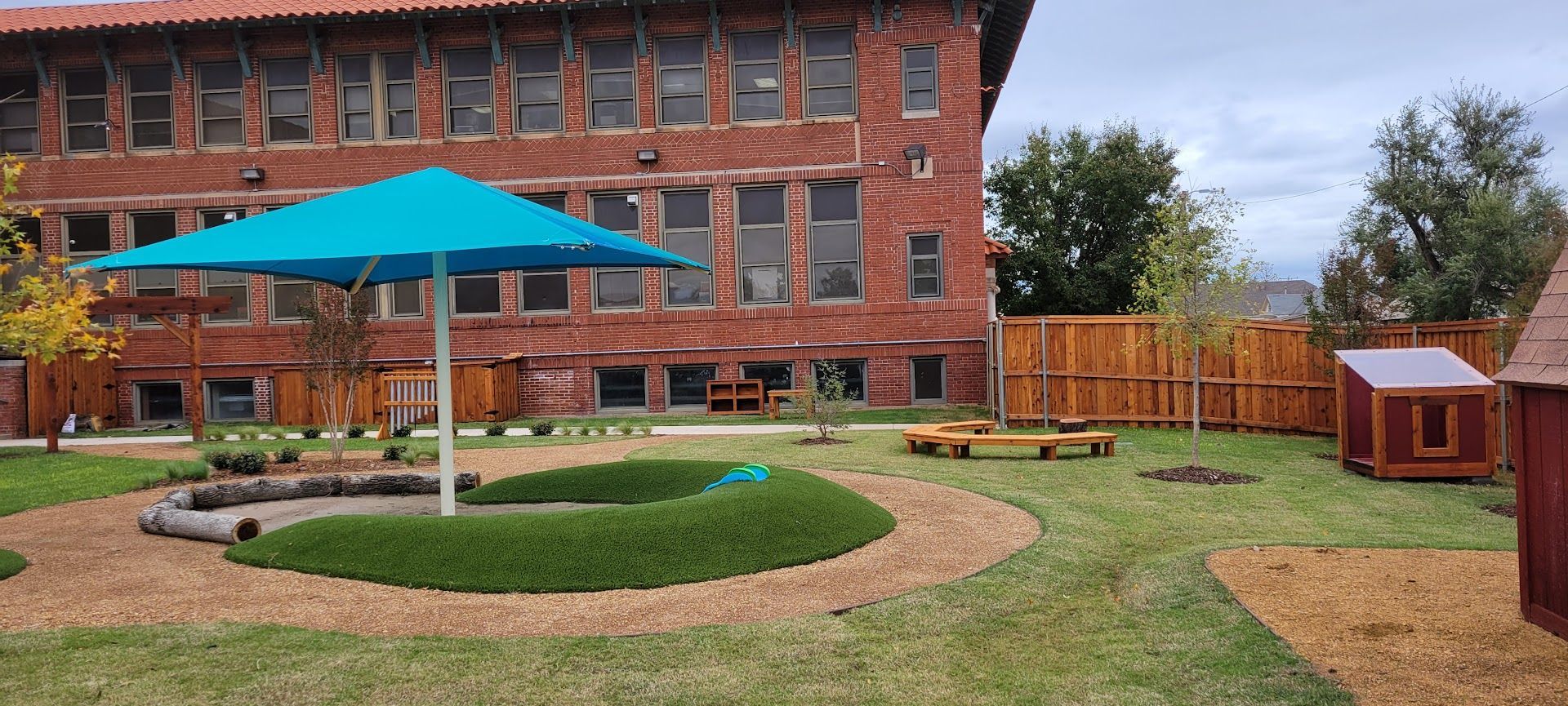 Brick courtyard with teal patio umbrella, circular stone garden, grass, and wooden bench by a building