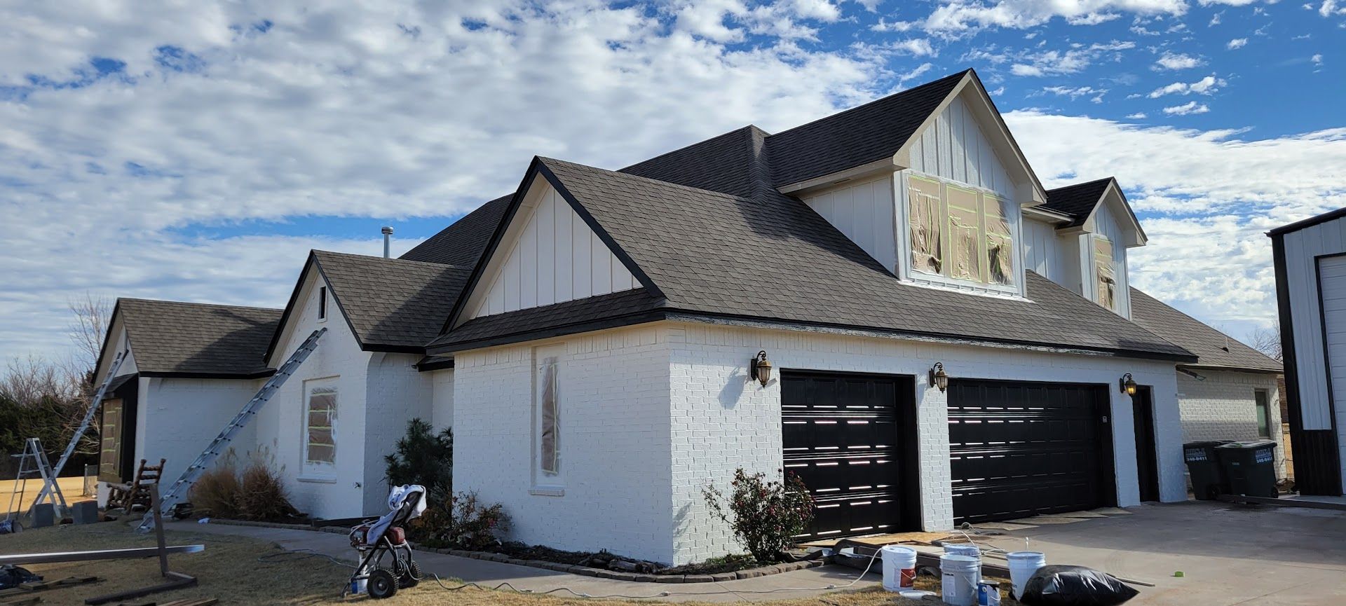 White house under construction with dark roof, black garage doors, and building materials outside.
