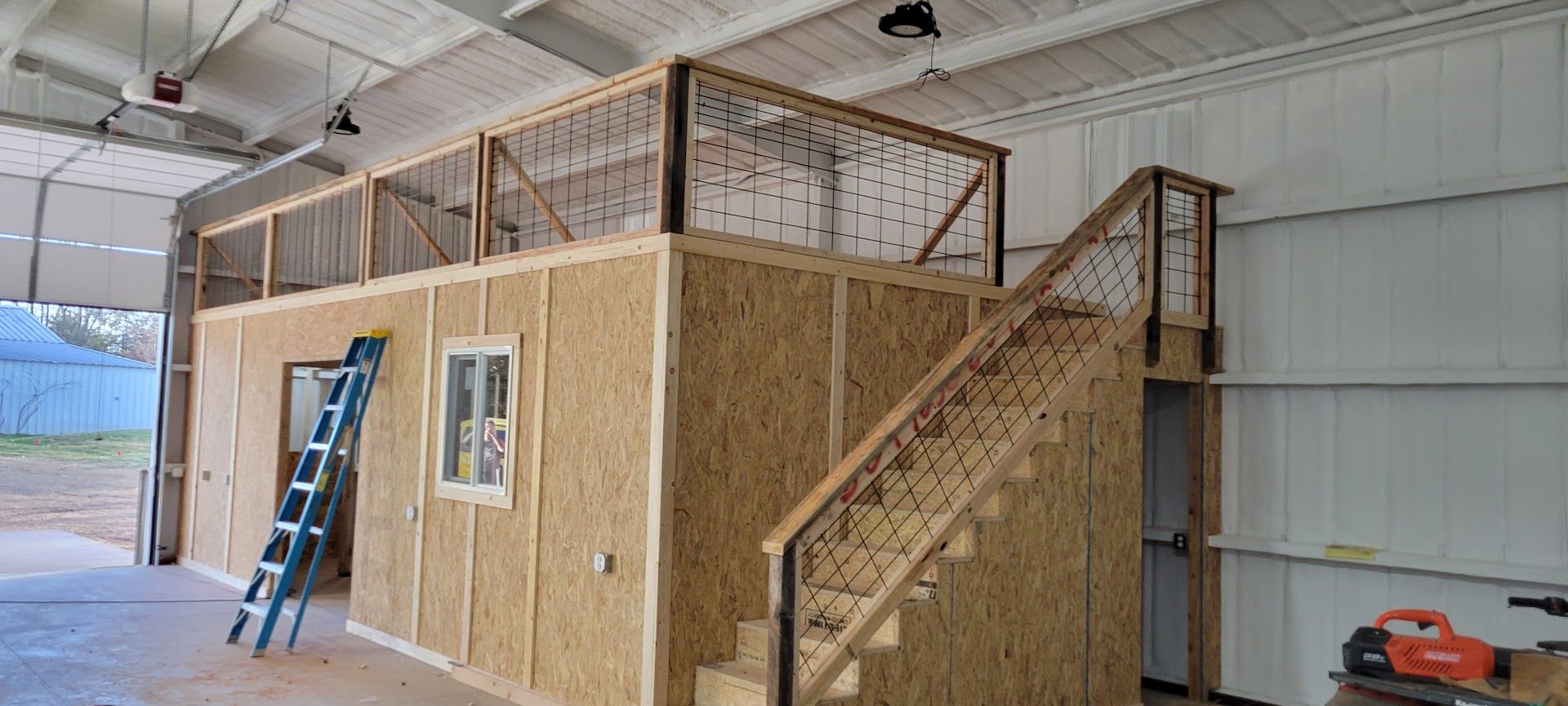 Interior of a large shed with a wooden mezzanine, stairs, ladder, and open garage door.