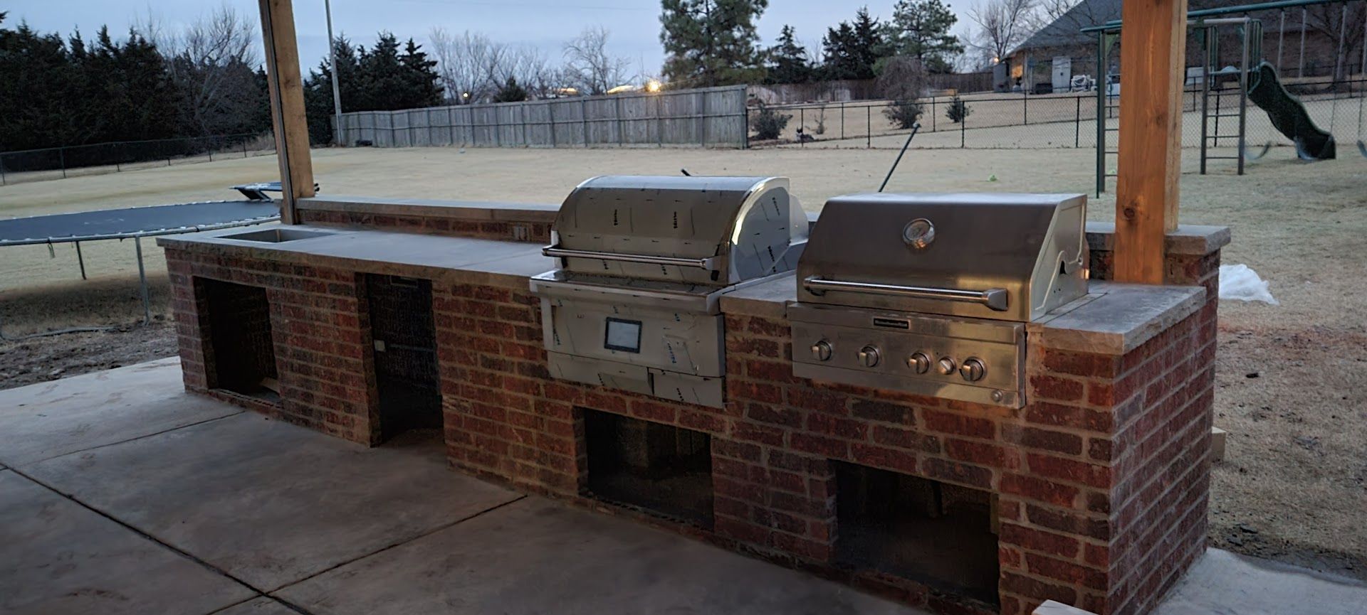 Outdoor brick barbecue grills under a pavilion in a park, with snow on the ground behind them.