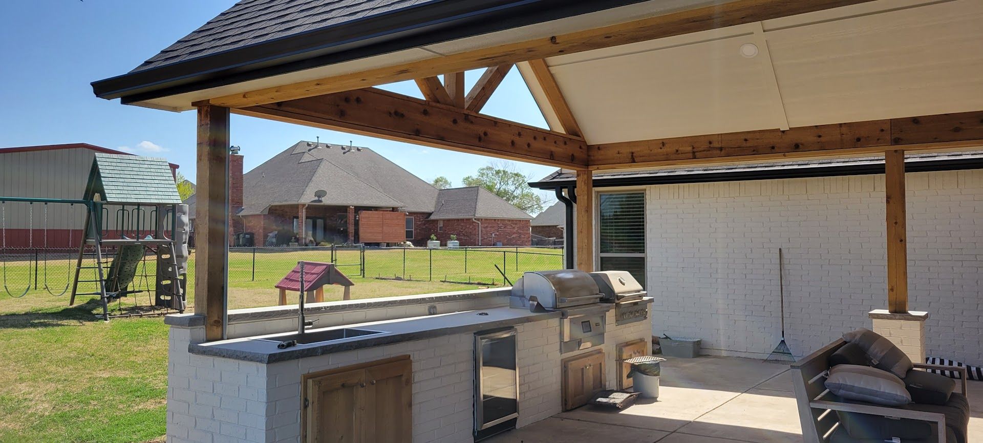 Outdoor patio with built-in grill and sink under a covered roof, overlooking a backyard and playground.