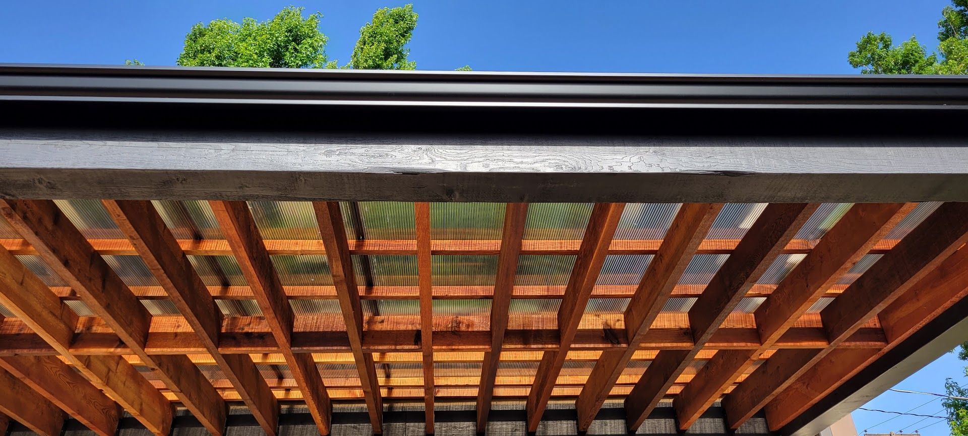 Wooden pergola roof with exposed beams against blue sky and green tree leaves