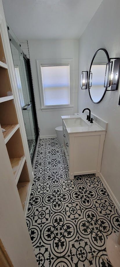 Narrow bathroom with patterned tile floor, white vanity, round mirror, and open doorway to a bright hall