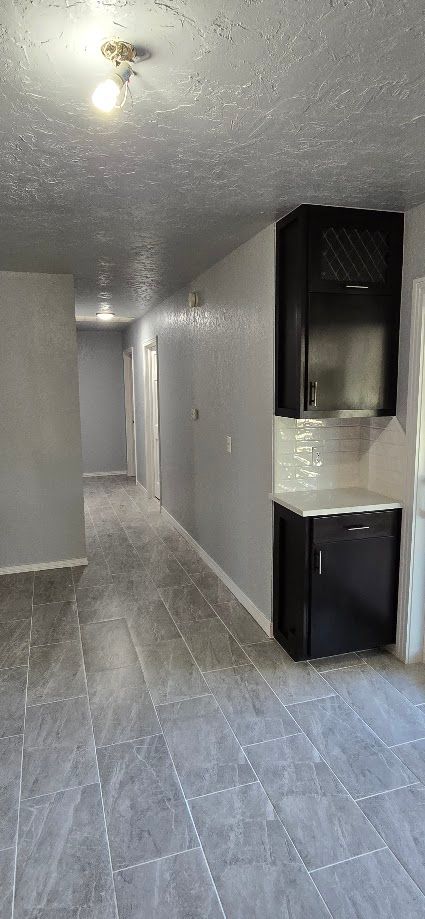 Narrow gray-tiled hallway with white walls, ceiling lights, and a small black kitchen counter on the right.