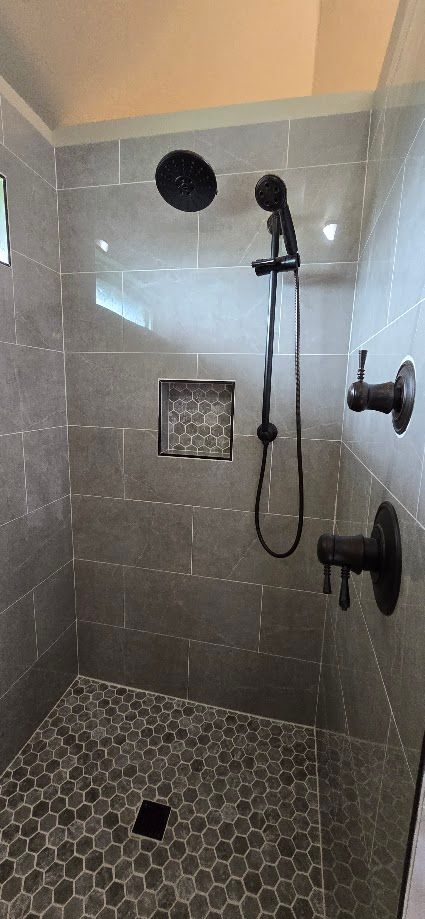 Small tiled shower with wall-mounted fixtures, showerhead, and pebble-style floor drain.
