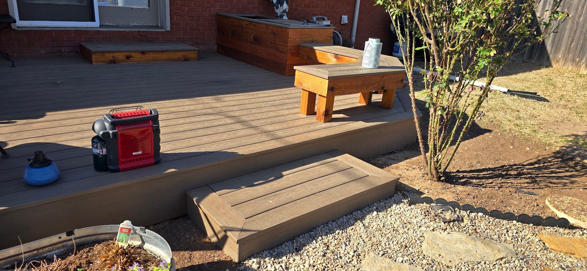 Wooden steps on a sunlit brick patio with a red toolbox, potted plant, and small tree nearby