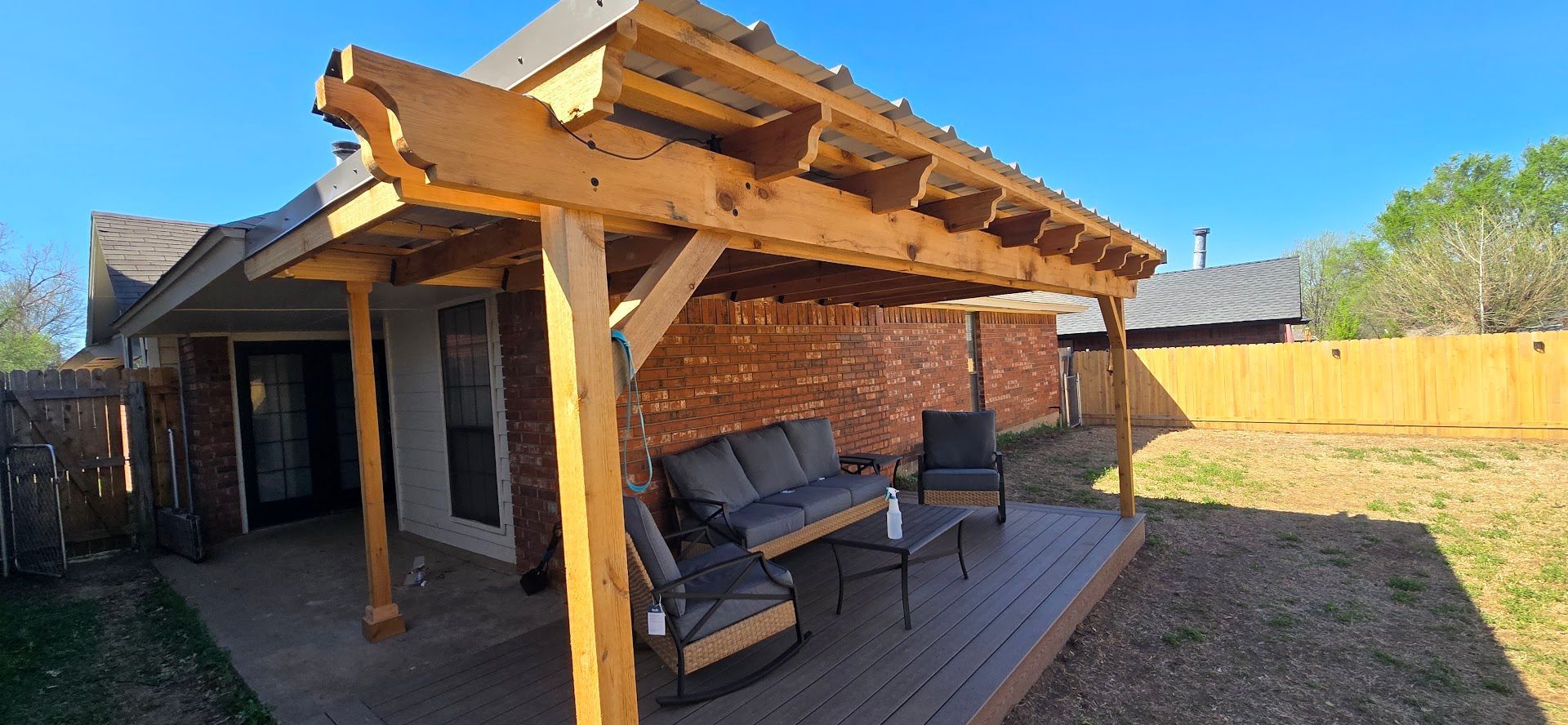 Backyard patio with a wooden pergola, seating, and a brick house wall in sunlight