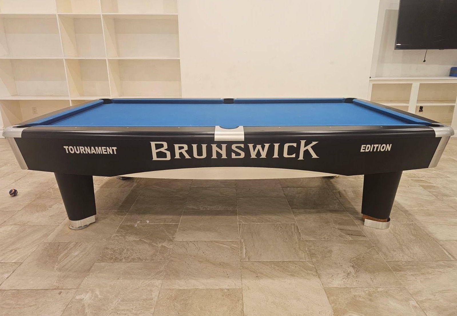 Brunswick pool table with blue felt in a bright room, centered against shelves and tiled floor