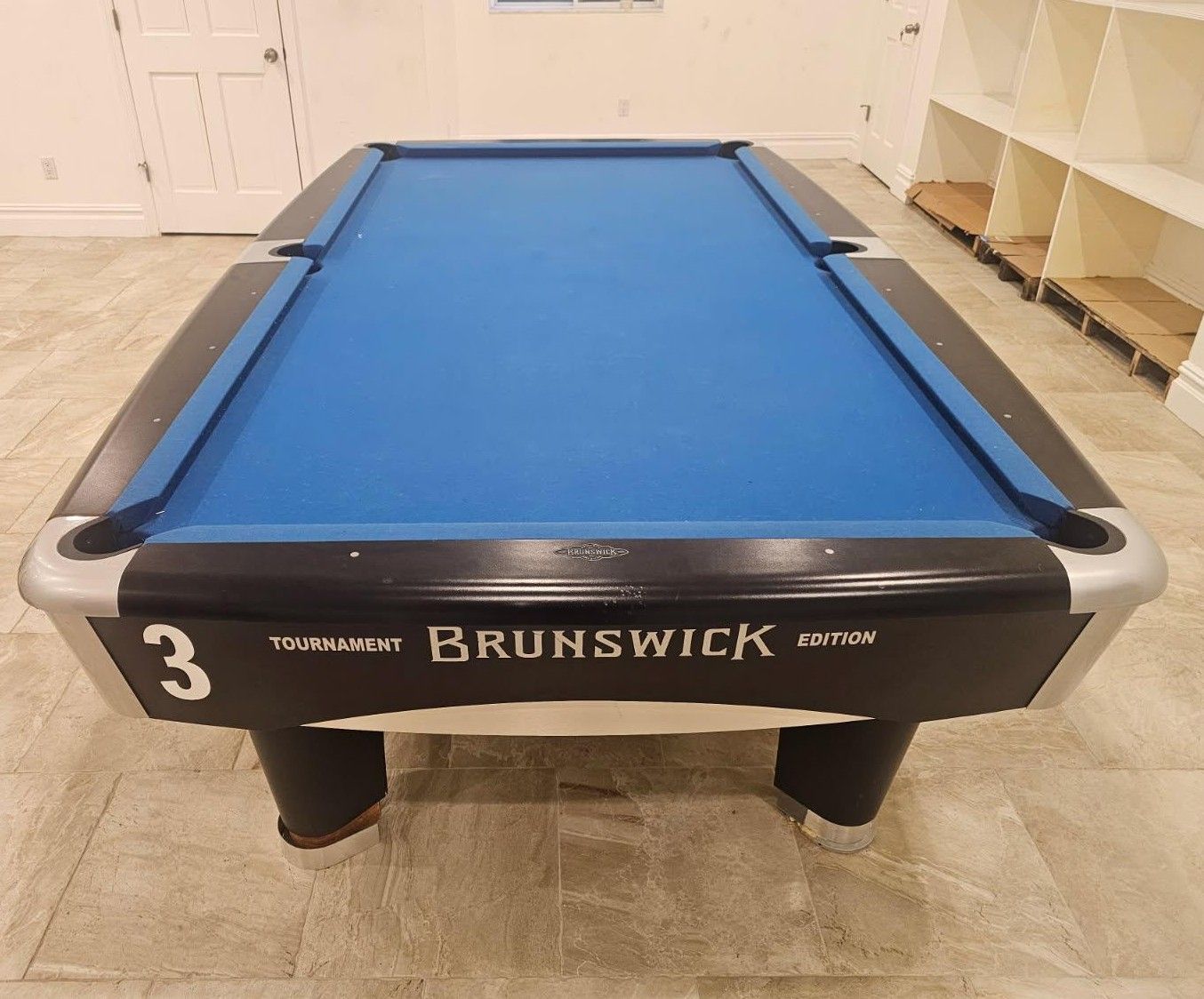 Blue Brunswick pool table in a room with light wood floor and shelving.