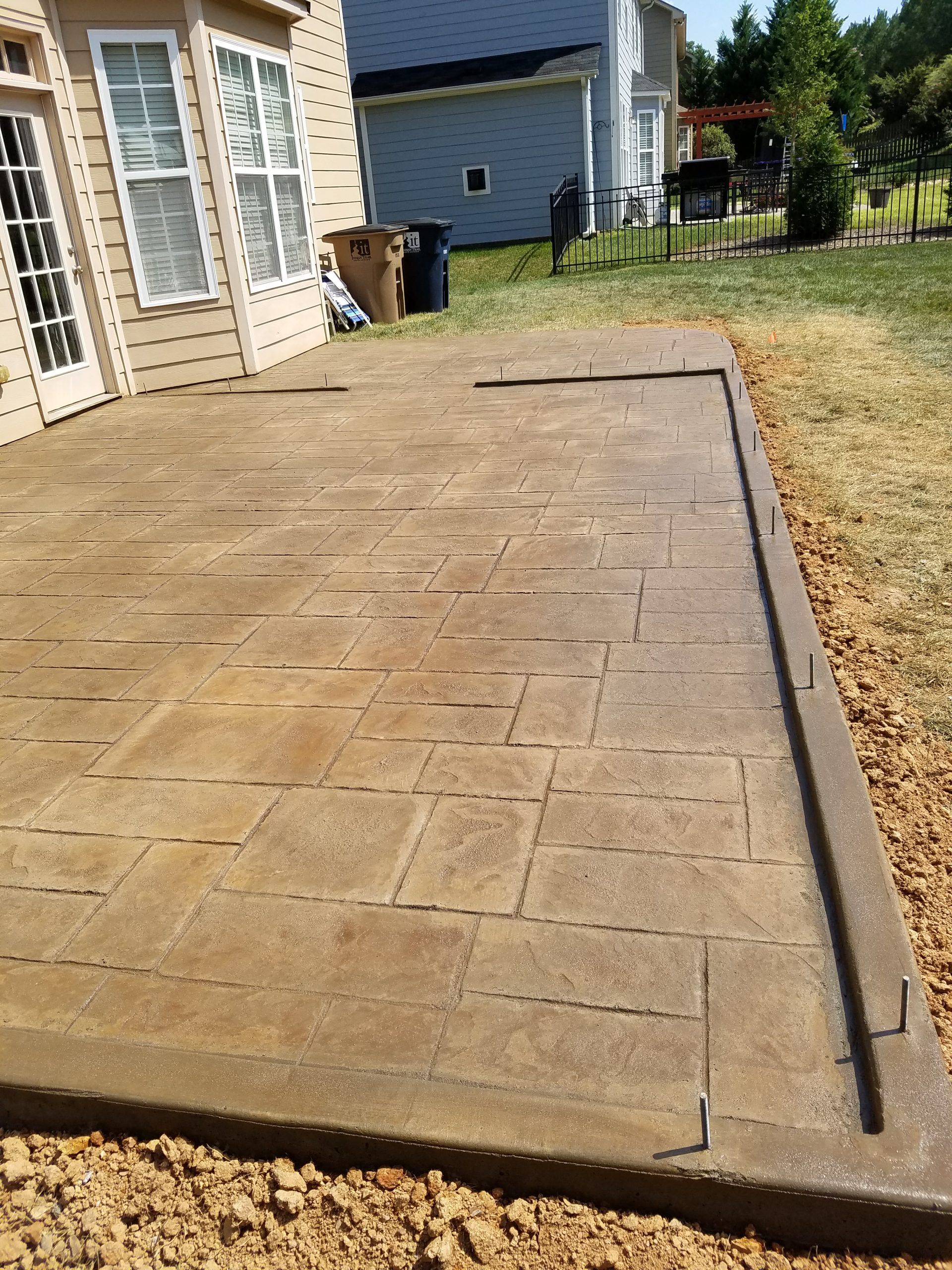 Concrete patio beside a house with a narrow border and dry grass yard in sunlight