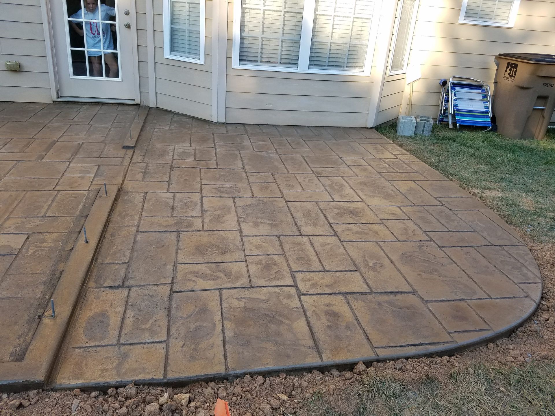 Stamped concrete patio beside a house, bordered by grass and a curved black edging strip.