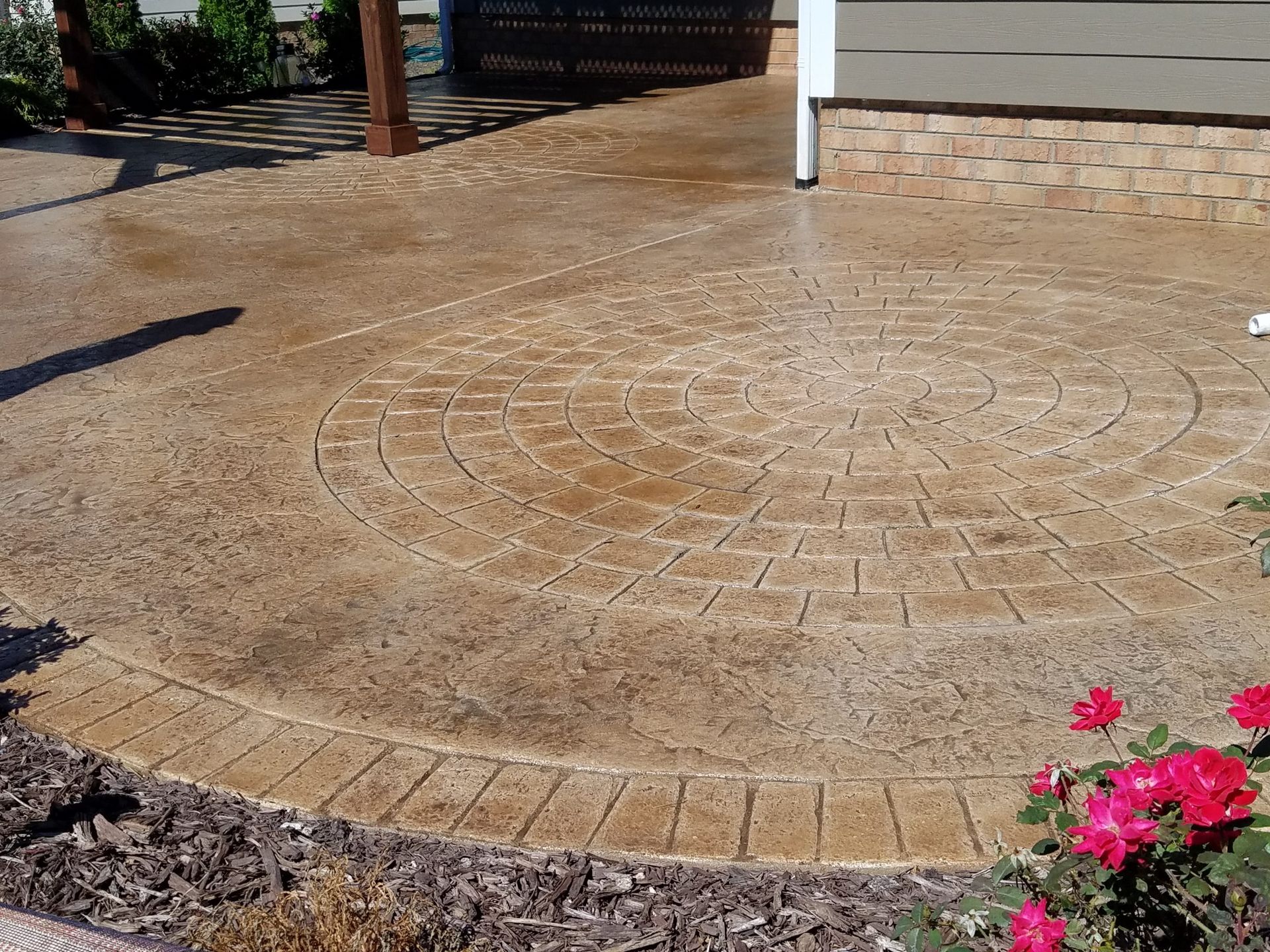 Stamped concrete patio with circular pattern next to a house, with pink flowers in the corner