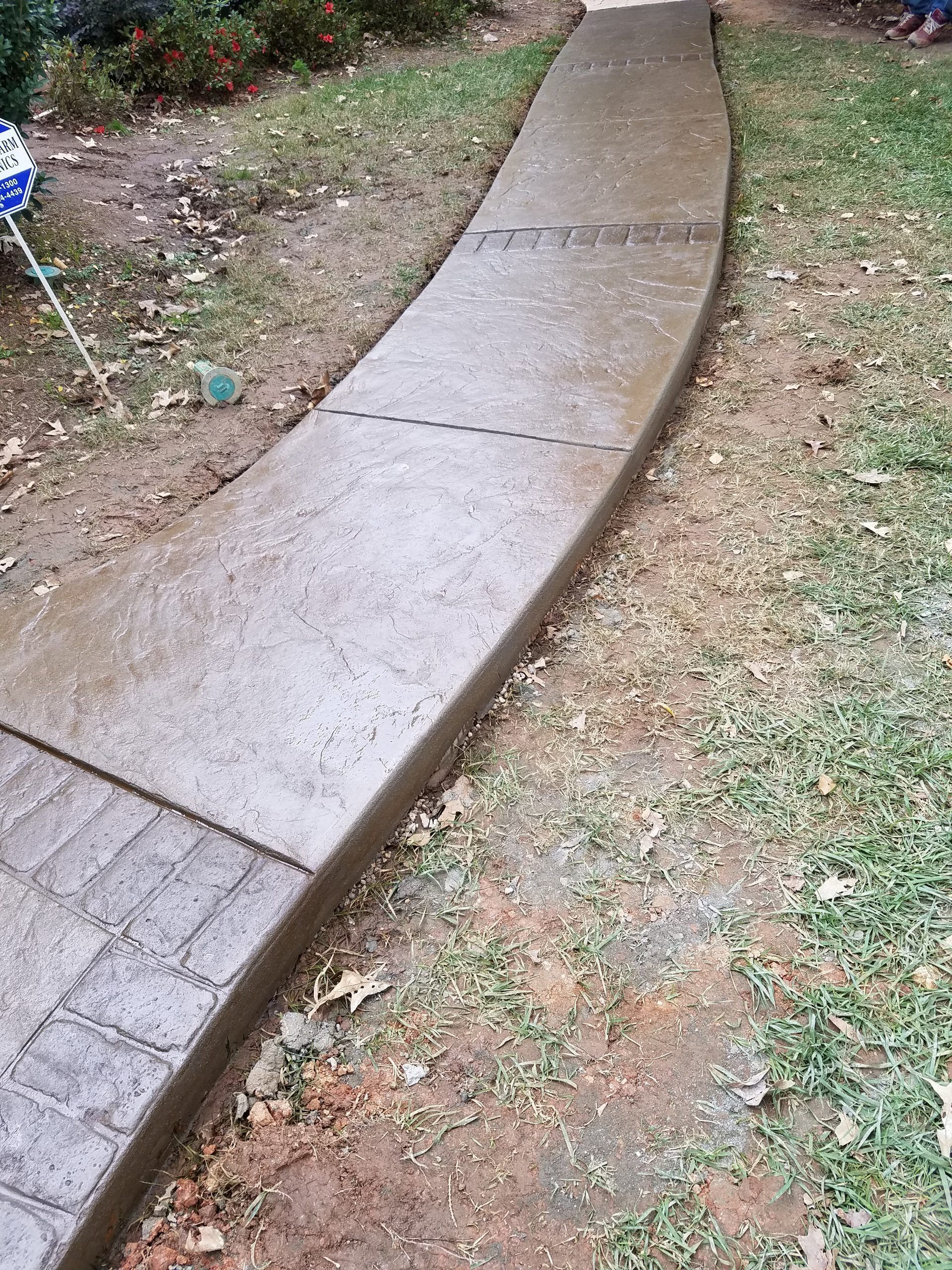 Curved concrete walkway bordered by grass and dirt, with a small brick section at the edge.