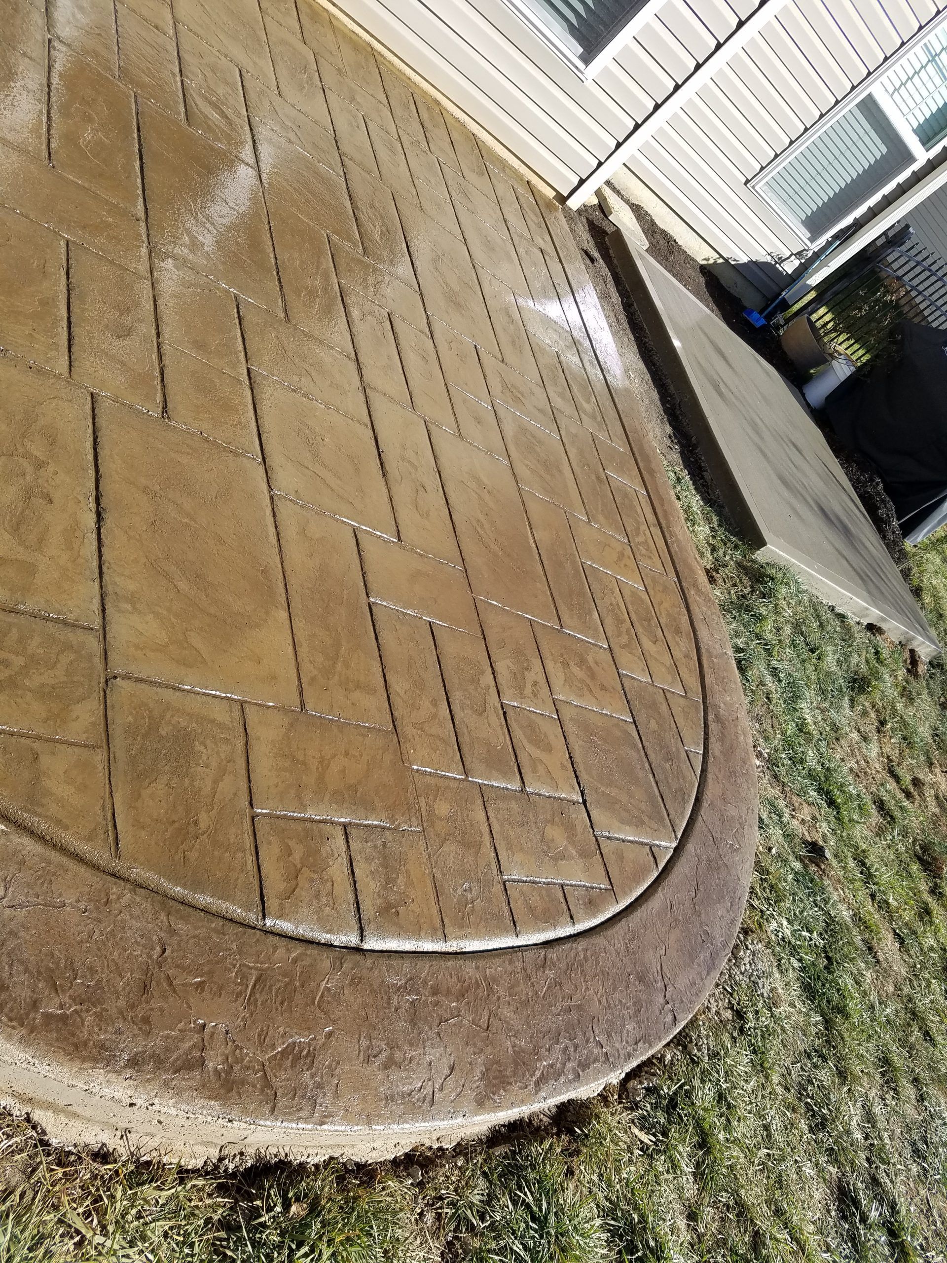 Wet stamped concrete patio beside a house, with a curved edge and a narrow garden border.
