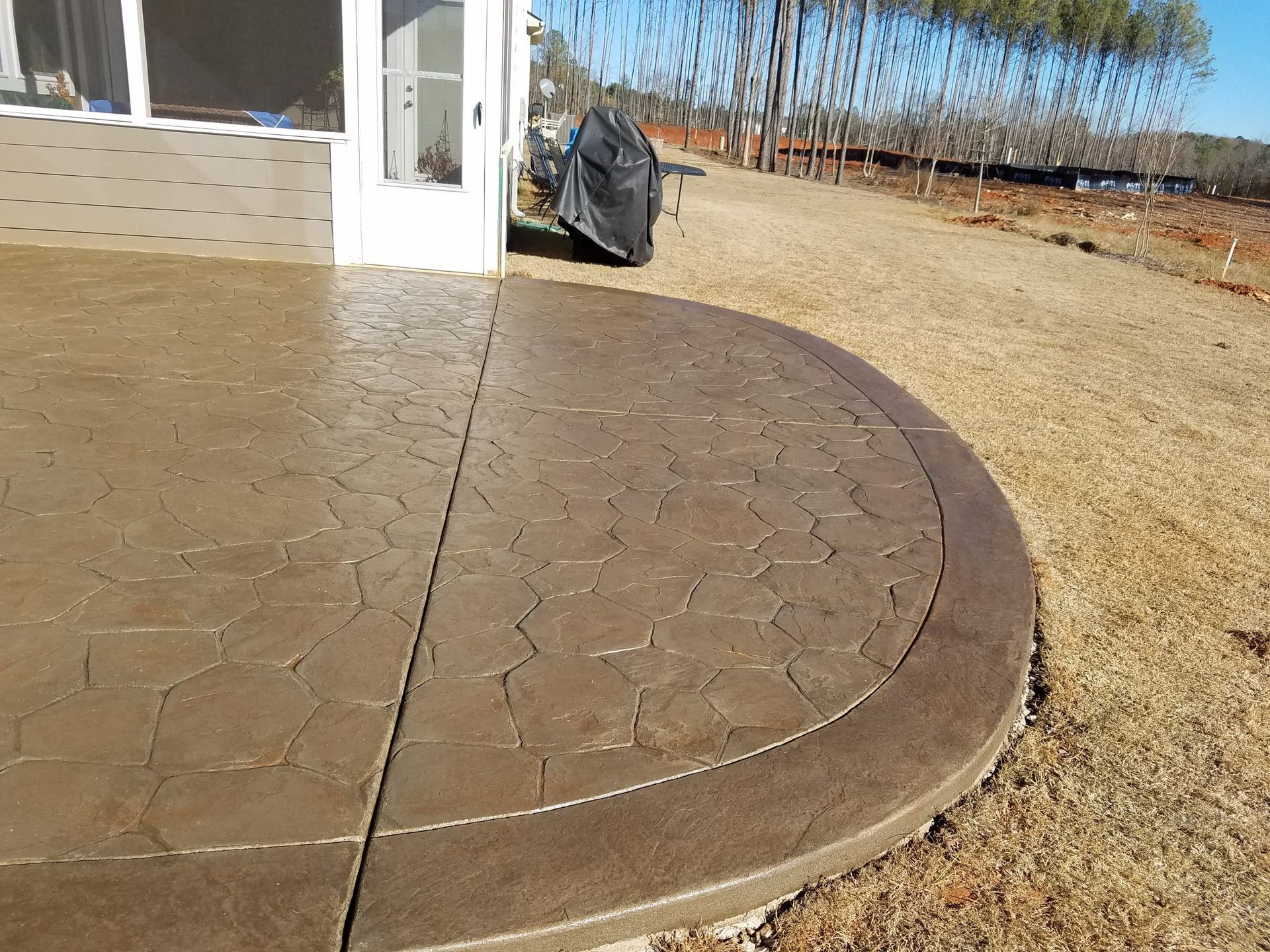 Curved concrete patio beside a house with a black grill and dry yard beyond