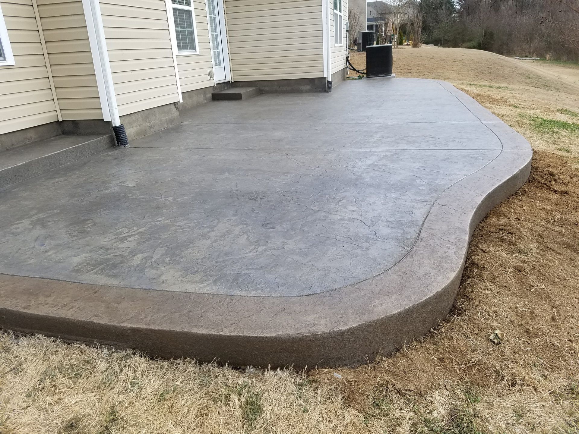 Fresh concrete patio slab beside a beige house, bordered by a curved edge and brown grass.