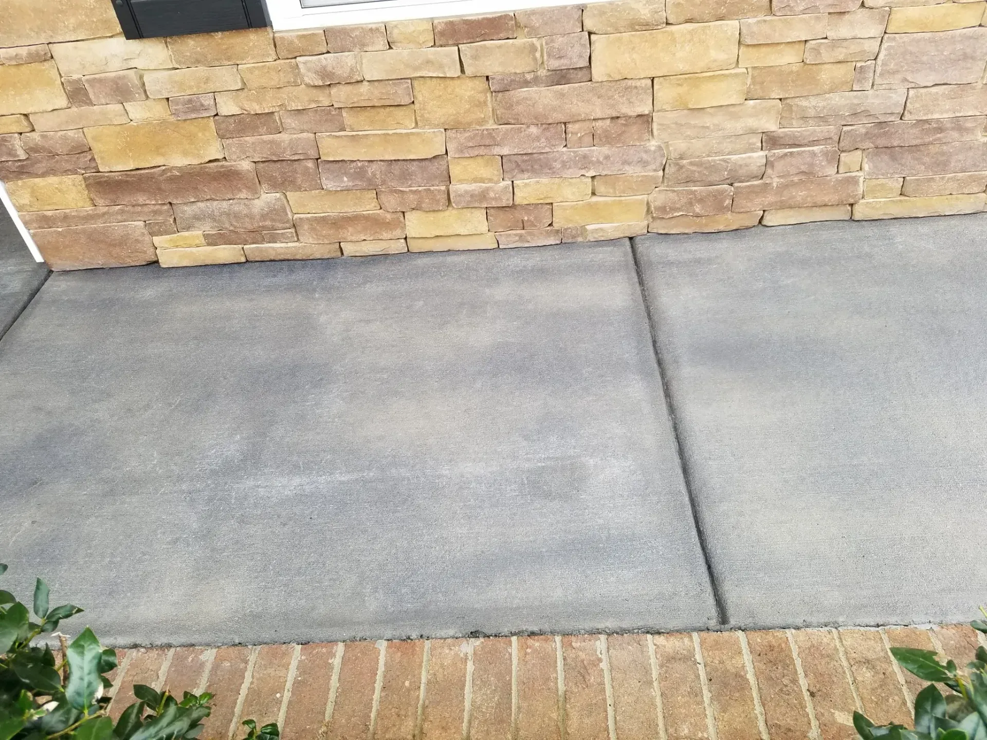 Concrete sidewalk beside a tan brick wall with a brick border and green plants at the lower left.