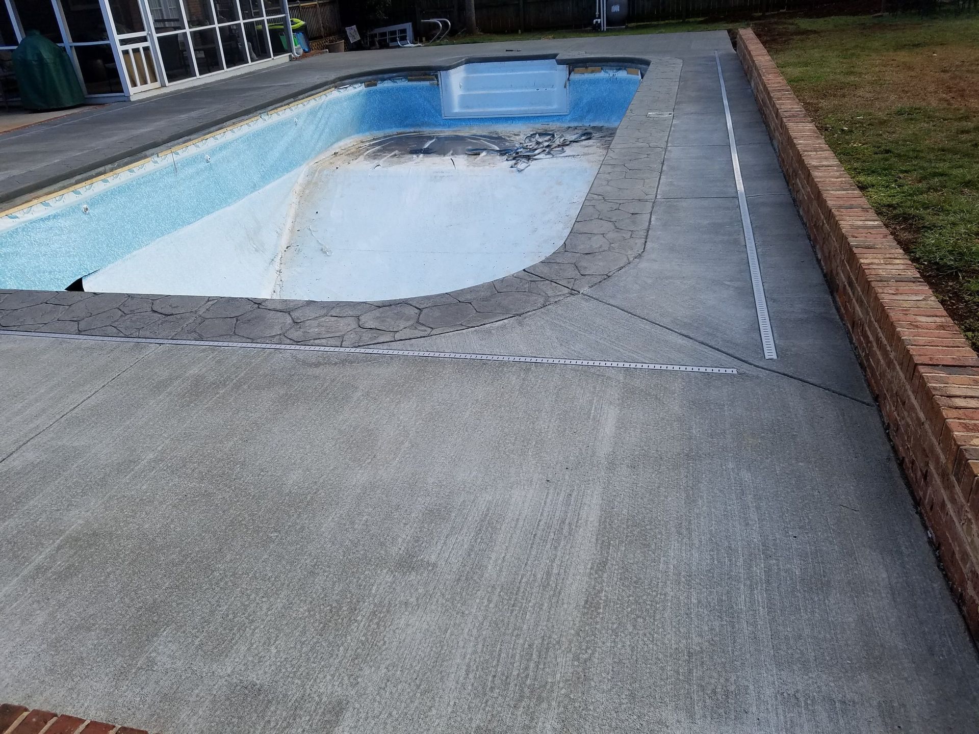 Empty outdoor swimming pool with cracked concrete deck and a brick border beside grass.