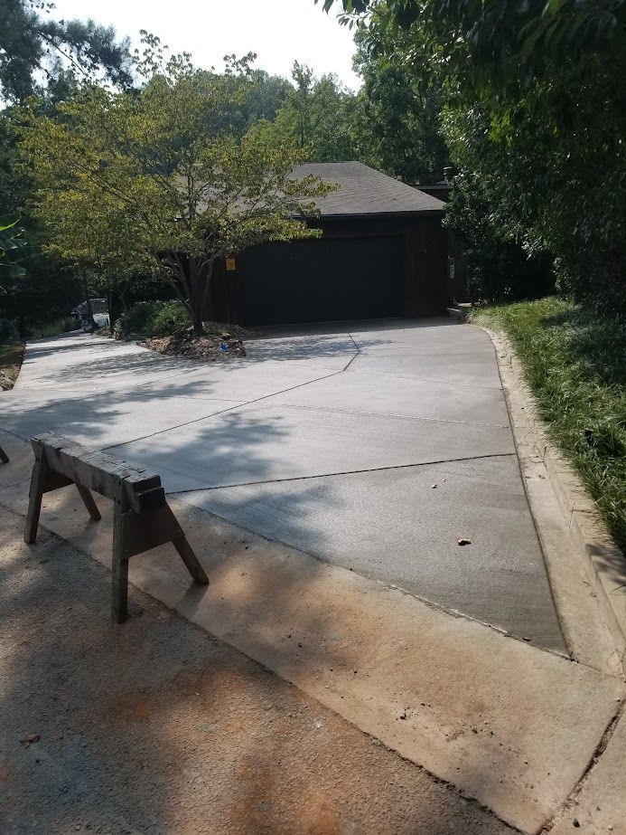 Concrete driveway leading to a garage, with trees and a wooden barrier in the foreground.