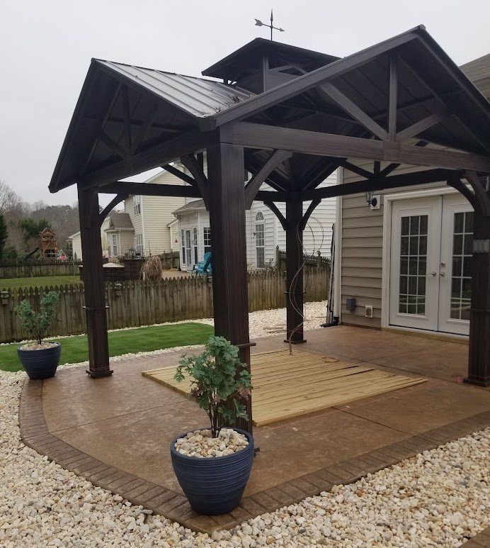 Covered wooden patio with dark beams, potted plants, and gravel border beside a house