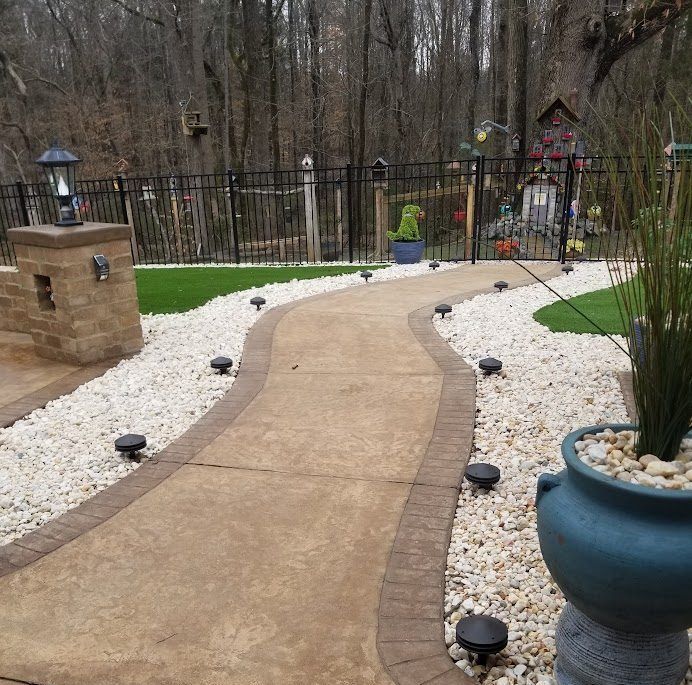 Curved concrete garden path bordered by white stones, with black lights and a blue planter beside a fenced yard.