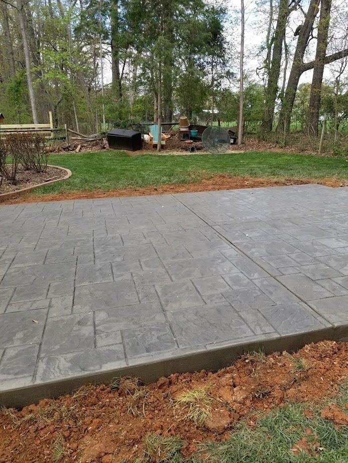 Gray stamped concrete patio in a wooded backyard with grass and trees beyond