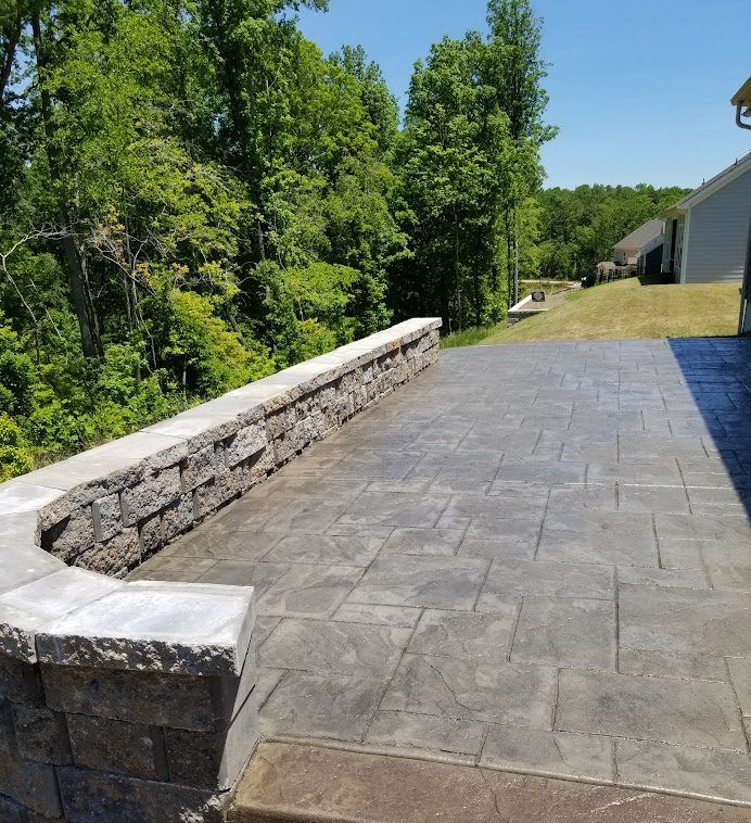 Stone patio with retaining wall beside a grassy yard and trees, under a clear blue sky