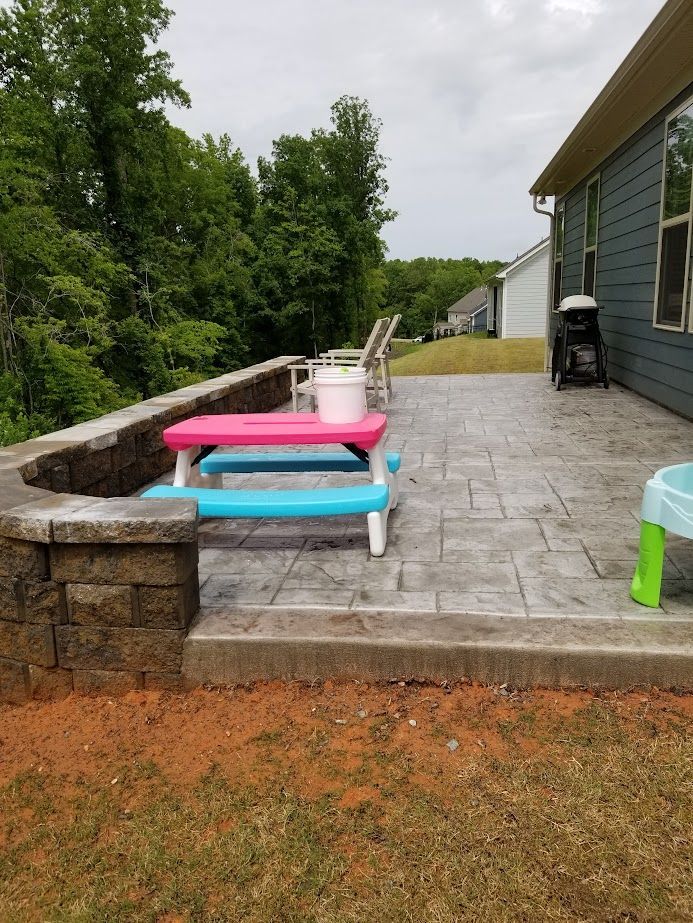 Stone patio beside a house with a bench, pink and blue table, and wooded yard.