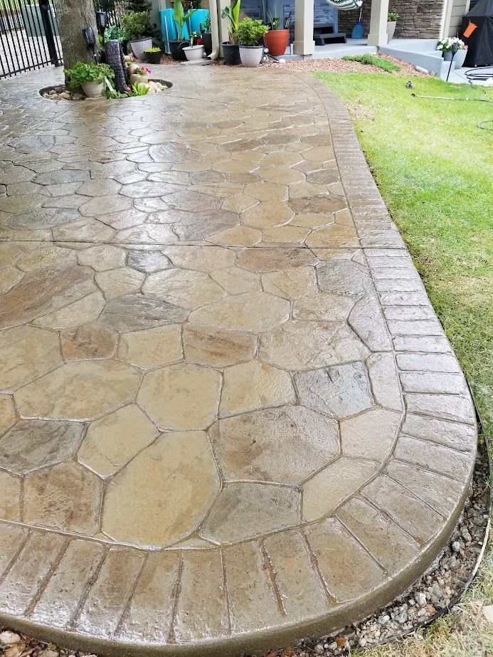 Curved stamped concrete patio with wet tan stones beside a strip of green lawn and potted plants