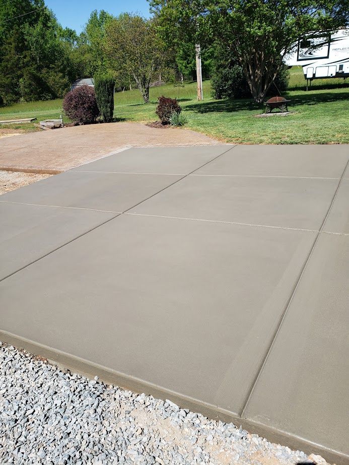 Freshly poured concrete driveway beside gravel, shrubs, and trees in a sunny yard