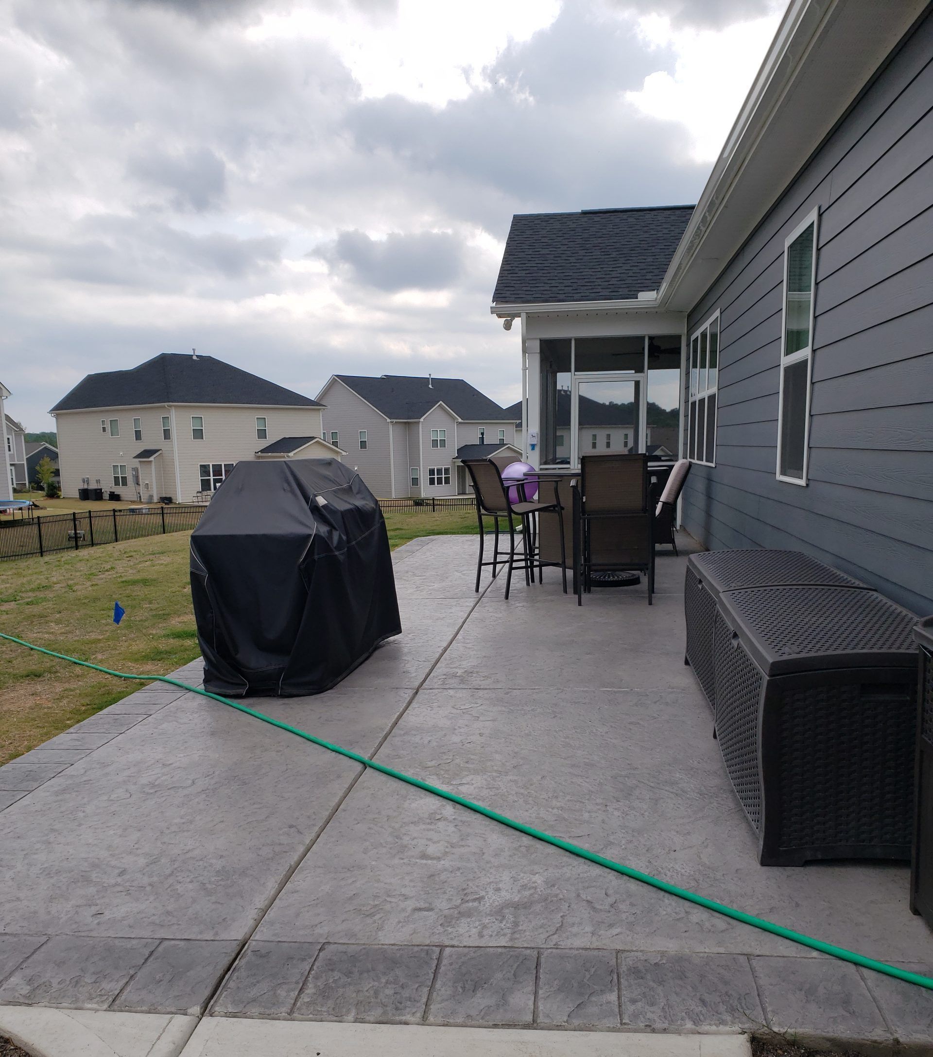 Back patio beside gray house with grill, patio furniture, and green garden hose on concrete.