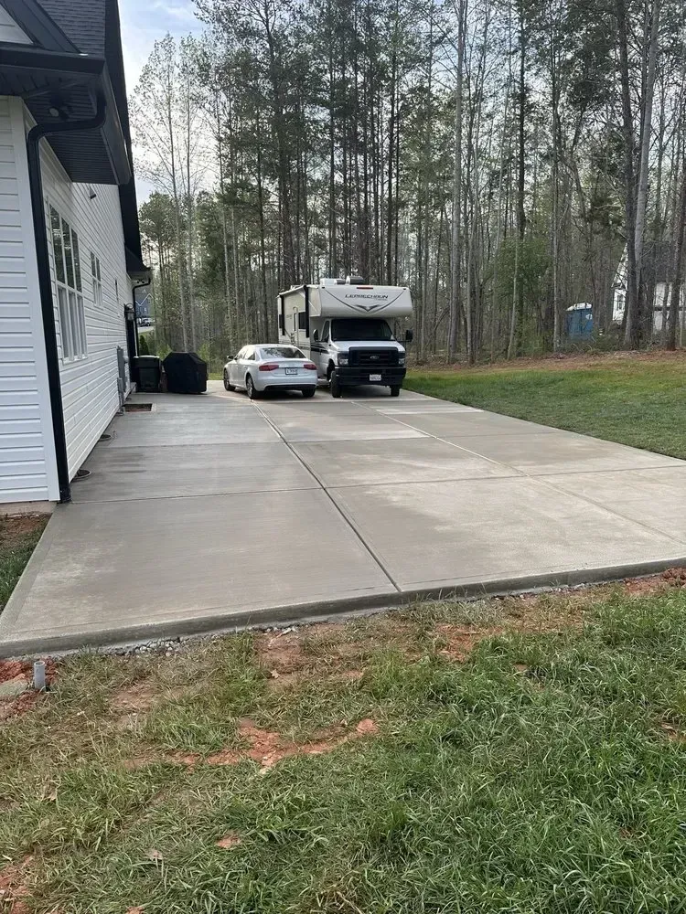Concrete driveway beside white house, with a parked car, utility truck, and wooded yard in the background.