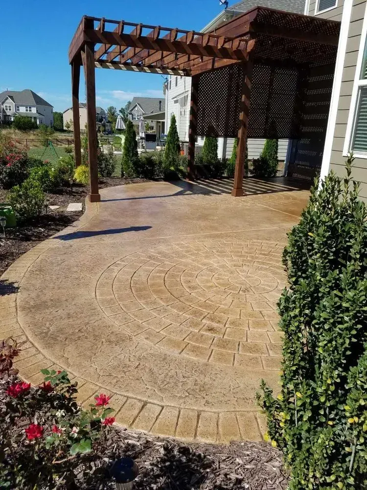 Backyard patio with patterned concrete, a wooden pergola, and landscaped garden beds under a blue sky