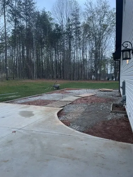 Wet concrete patio beside house, curving walkway, and wooded backyard on a rainy day