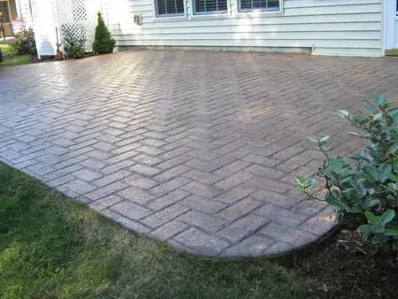 Brick patio beside a house, with a curved edge and surrounding grass and shrubs